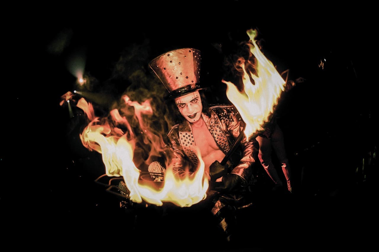 Fire performer wearing a top hat and costume surrounded by flames on a dark stage.